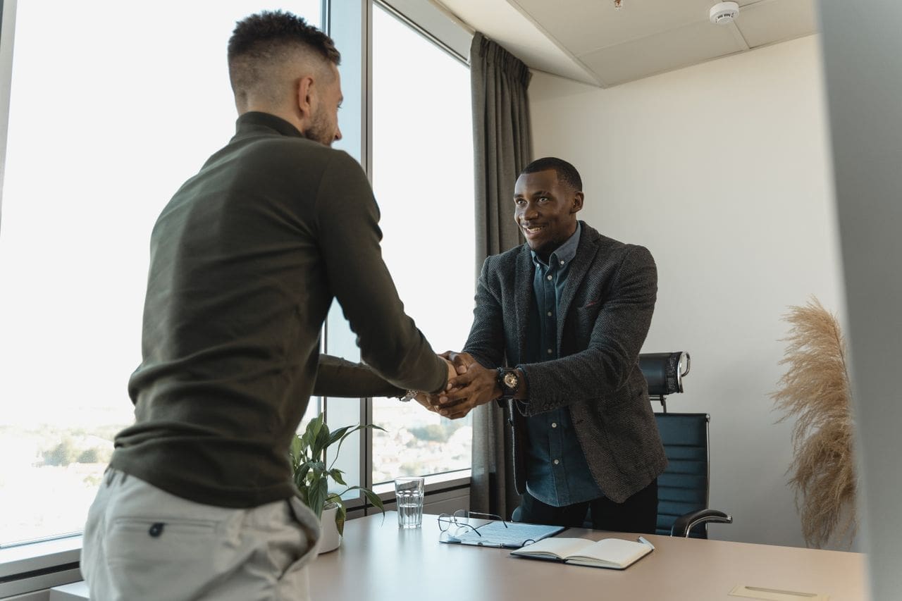 why-choose-us Two businessmen exchanging a handshake during a professional meeting in a modern office setting.